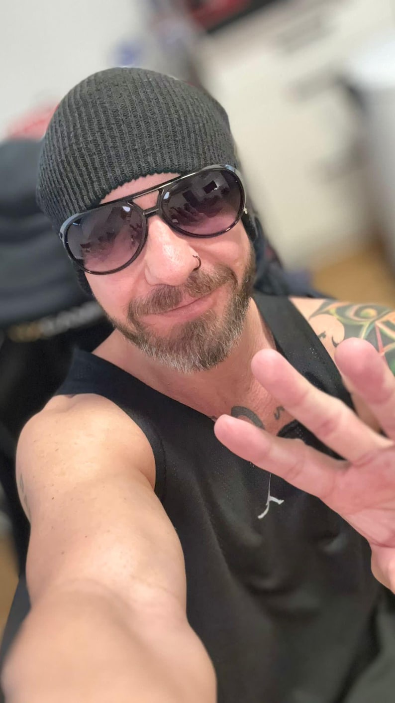 May include: A person wearing a black beanie and sunglasses with a dark frame. The person is also wearing a black tank top. The person is holding up their hand with three fingers extended.