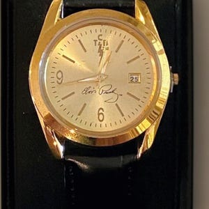 以下が含まれることがあります： 黒い革バンドが付いた金色の腕時計。文字盤は金色で、黒い数字と針が付いています。日付は3時の位置に表示されます。「Elvis Presley」の筆記体で刻印されています。黒い面に置かれた時計。