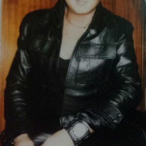 May include: A person wearing a black leather jacket, black pants, and black boots. The person is also wearing a watch and sunglasses. The background is a wooden door.