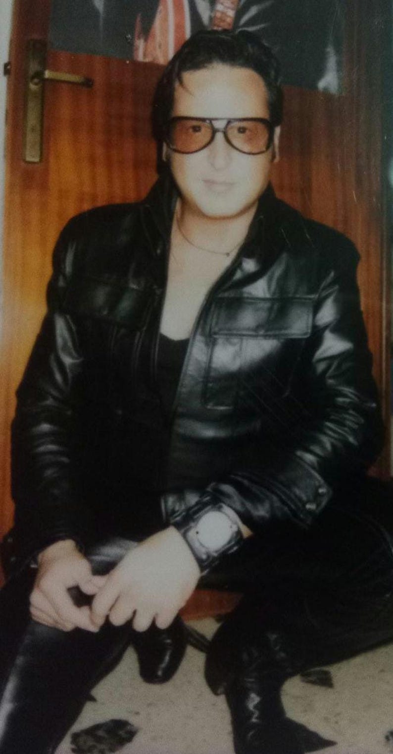 May include: A person wearing a black leather jacket, black pants, and black boots. The person is also wearing a watch and sunglasses. The background is a wooden door.