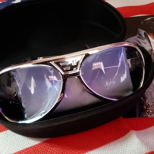 Puede incluir: Gafas de sol con montura plateada y lentes azules en un estuche negro abierto. Las gafas de sol tienen un estilo aviador distintivo. Una imagen de un cantante con un atuendo con lentejuelas es visible en el fondo, sobre una bandera tricolor.
