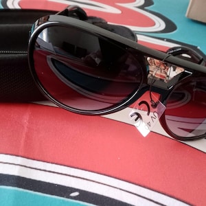 May include: Black sunglasses with dark tinted lenses and a thick frame. The sunglasses have a decorative emblem on the upper corner of the frame. A black case is visible in the background. A tag with "CE UV 400" is attached.