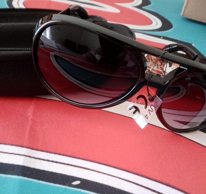May include: Black sunglasses with dark tinted lenses and a thick frame. The sunglasses have a decorative emblem on the upper corner of the frame. A black case is visible in the background. A tag with "CE UV 400" is attached.
