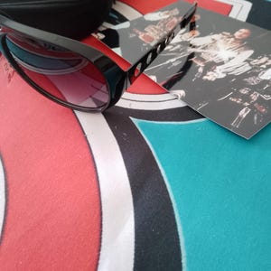 May include: Black sunglasses with a black case and a photo card. The sunglasses have a thick frame and red-tinted lenses. The case is rectangular and has a snap closure. The photo card has a black and white image.