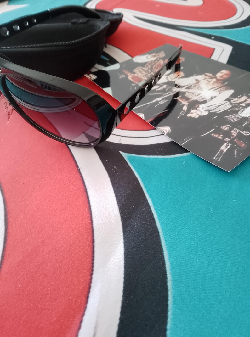 May include: Black sunglasses with a black case and a photo card. The sunglasses have a thick frame and red-tinted lenses. The case is rectangular and has a snap closure. The photo card has a black and white image.