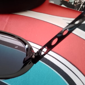 May include: Black sunglasses with oval frames and dark lenses. The temples have a unique design with elongated cutouts. A black case and a card with a picture are in the background. The sunglasses are on a red, white, and blue patterned surface.