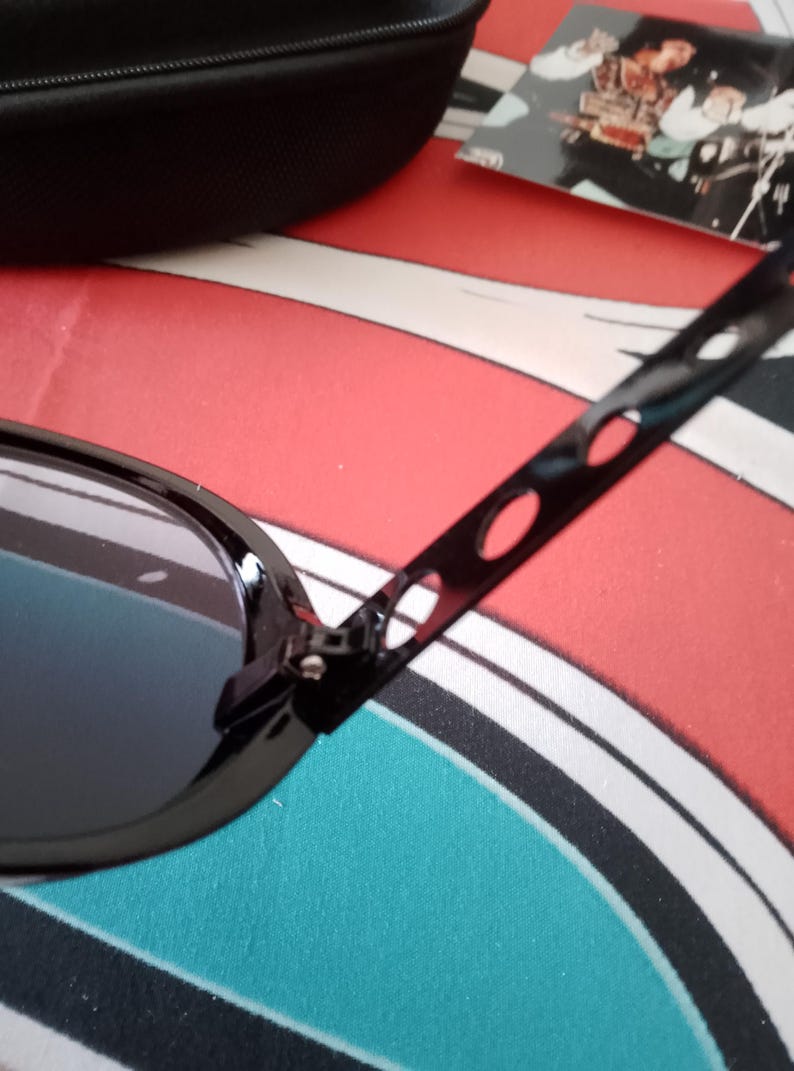 May include: Black sunglasses with oval frames and dark lenses. The temples have a unique design with elongated cutouts. A black case and a card with a picture are in the background. The sunglasses are on a red, white, and blue patterned surface.