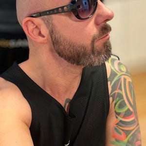 May include: Black sunglasses with a dark lens and a black frame with small circular cutouts. The person is wearing a black tank top. The person has a shaved head and a beard.