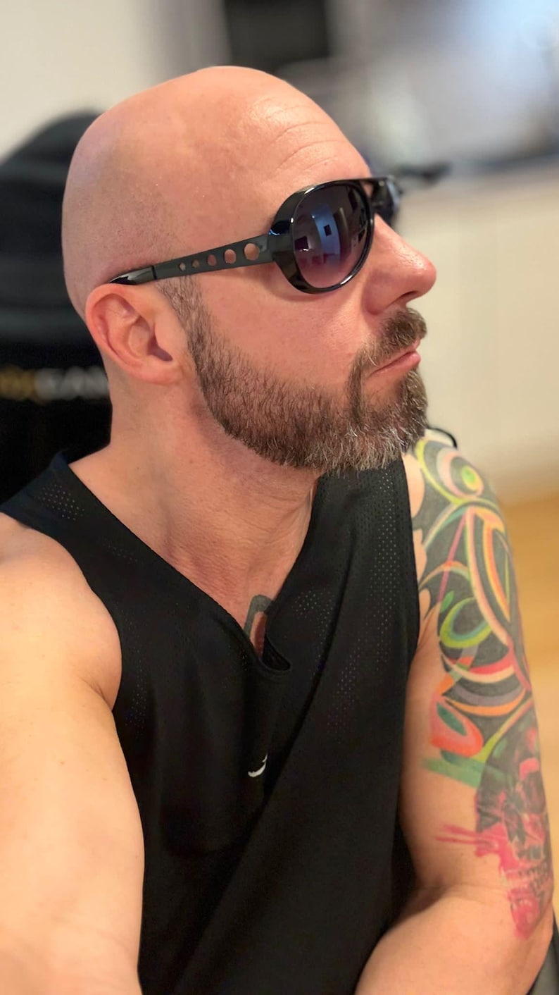 May include: Black sunglasses with a dark lens and a black frame with small circular cutouts. The person is wearing a black tank top. The person has a shaved head and a beard.