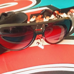 May include: Black sunglasses with red-tinted lenses and a black case. The sunglasses have a thick frame and a small red accent on the bridge. A small tag with the text "UV 400" is attached to the frame.