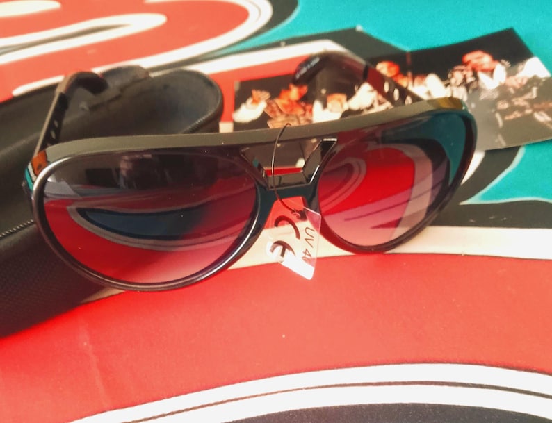 May include: Black sunglasses with red-tinted lenses and a black case. The sunglasses have a thick frame and a small red accent on the bridge. A small tag with the text "UV 400" is attached to the frame.