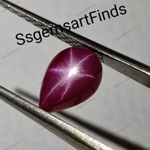 Peut inclure: Une pierre précieuse rouge foncé en forme de poire, présentant un motif en étoile à six rayons. La surface polie reflète la lumière, créant un effet d'étoile. La gemme est tenue par une pince métallique. Le texte "SsgemsartFinds" est visible.