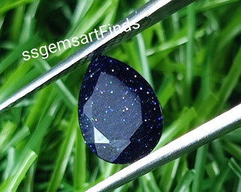 May include: A teardrop-shaped blue gemstone with a sparkly, glittery finish. The gemstone is held by tweezers against a green grassy background.