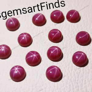 May include: A collection of round, cabochon-cut ruby gemstones. Each gemstone displays a six-rayed star effect, with white lines radiating from the center. The rubies are a deep, rich red color and are set against a light background. The text "SsgemsartFinds" is in the top left corner.