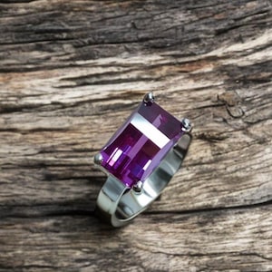 Peut inclure: Une bague en argent ornée d'une grande pierre précieuse rectangulaire violette. La pierre précieuse à facettes reflète la lumière, sertie sur un simple anneau en argent. La bague est posée sur une surface en bois texturée.