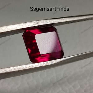 Può includere: Una pietra preziosa di colore rosso intenso con forma quadrata, tenuta da una pinzetta. La pietra preziosa è tagliata con sfaccettature e ha un'alta lucentezza.