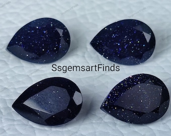 May include: Four pear-shaped blue gemstone cabochons with a sparkly, speckled texture.  The gemstones are arranged in a square pattern on a white surface.  Ssgemsartfinds.