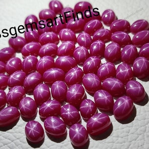 May include: A collection of 40 pink star ruby cabochons. The stones are oval shaped and have a star-like pattern in the centre. The stones are all approximately the same size and are arranged in a random pattern.