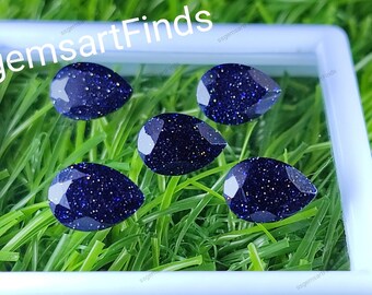 May include: Five pear-shaped blue gemstone cabochons with a sparkly, speckled texture. The stones are displayed in a white square box on a green grassy background.