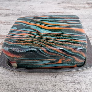 Könnte beinhalten: Eine quadratische Butterdose aus Keramik mit einer bunten, marmorierten Glasur in Grün-, Blau-, Orange- und Weißtönen. Die Dose steht auf einem dunkelgrauen Tablett.