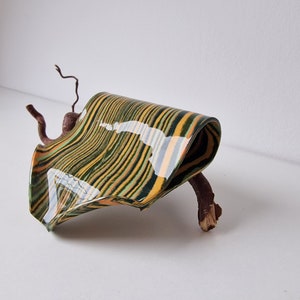 Könnte beinhalten: Eine dekorative Skulptur aus Holz mit einem grün-gelb-orangen Streifenmuster. Die Skulptur ist wellenförmig und auf einem kleinen Ast montiert.