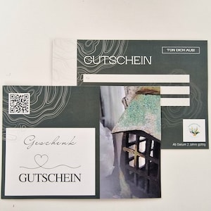 Könnte beinhalten: Ein grüner Geschenkgutschein mit weißem Rand und dem Text "Gutschein" in Schwarz. Der Gutschein hat einen QR-Code und ein kleines Bild eines Fensters mit einem Kreuz.