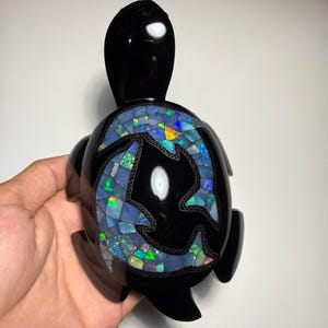 Puede incluir: Una figura de tortuga negra pulida con una incrustación decorativa en el caparazón. El caparazón muestra dos siluetas de delfines rellenas con un mosaico de fragmentos de ópalo iridiscentes y multicolores que brillan con reflejos azules, verdes y naranjas.
