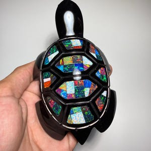 Puede incluir: Una figura de tortuga negra con un caparazón incrustado con segmentos de ópalo iridiscentes y multicolores. La tortuga tiene un cuerpo negro liso y pulido con un acento blanco en la cabeza. El caparazón está compuesto por secciones geométricas rellenas de piezas de ópalo vibrantes en tonos azules, verdes, rojos y naranjas.