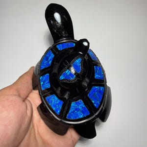 Puede incluir: Una figura de tortuga negra con acabado pulido y segmentos de piedra azul iridiscente incrustados en el caparazón. El centro del caparazón muestra un motivo de mariposa grabado relleno de piedra azul a juego, creando un contraste vivo.