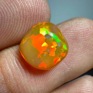 Puede incluir: Una piedra preciosa de ópalo pulida, de forma ovalada, con vibrantes patrones internos naranjas, amarillos y verdes. El ópalo se sostiene en la mano de una persona, mostrando sus colores naturales y su estructura interna única. El ópalo es natural y sin tratar.