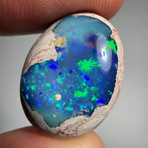 Pode incluir: Uma pedra preciosa de opala boulder de formato oval com um centro azul profundo e translúcido com flashes vibrantes de verde neon e azul elétrico, cercada por uma matriz rochosa porosa, texturizada e clara com veios castanho-avermelhados.