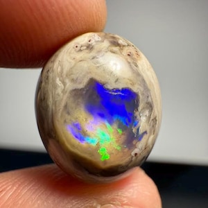 Op de afbeelding: Een gepolijste, ovaalvormige opaal edelsteen. De opaal heeft een crèmewitte en bruine buitenkant met een levendige weergave van interne kleuren, waaronder blauw, groen en geel. De edelsteen wordt tussen twee vingers gehouden.