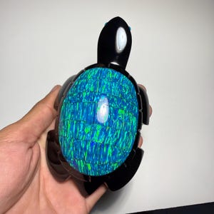 Puede incluir: Figura de tortuga negra con caparazón de mosaico azul y verde. La cabeza y las aletas de la tortuga son negras, y el caparazón tiene un aspecto brillante y texturizado. La figura se sostiene en una mano sobre un fondo blanco.