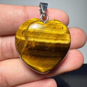 Puede incluir: Colgante en forma de corazón de ojo de tigre. La piedra pulida tiene un color marrón dorado con bandas marrones oscuras. Tiene una fianza de metal plateado para una cadena. La piedra es lisa, mostrando patrones naturales. Un colgante único.