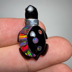 Puede incluir: Colgante artesanal con forma de tortuga. El cuerpo negro presenta detalles de ópalo iridiscente y una banda de metal plateado. El ópalo muestra una vibrante gama de colores, incluyendo rojo, naranja, amarillo y verde. Una joya única.
