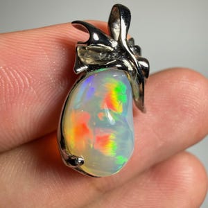 Puede incluir: Un colgante de ópalo en forma de lágrima engastado en un marco de metal plateado pulido. La piedra preciosa muestra una base blanca translúcida con destellos vibrantes de rojo, naranja y verde en toda su superficie lisa y redondeada.