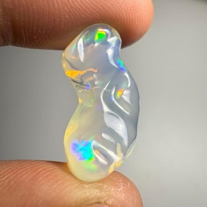 以下が含まれることがあります： 緑、青、黄色の虹色の閃光を持つ、半透明の洋梨型のオパール宝石。 研磨された石は滑らかで光沢のある表面を持ち、2本の指の間に保持されています。 オパールの色は光によって変化し、色の遊びを作り出します。