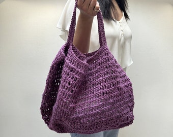 May include: A purple crocheted tote bag with a wide, open top and a single handle. The bag is made of a thick, textured yarn and has a loose, open weave.