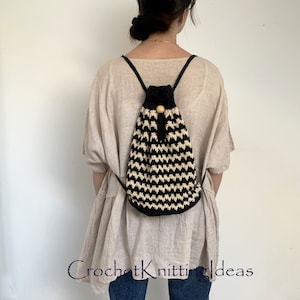 以下が含まれることがあります： 木製の留め具が付いた、黒と白のクロシェの巾着バックパック。バックパックは人が背負っています。