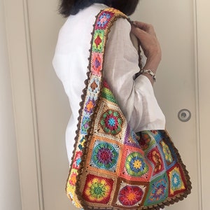 Può includere: Una borsa a mano in crochet colorata con una tracolla marrone. La borsa è realizzata con quadrati con diversi motivi floreali in tonalità di rosso, arancione, giallo, verde, blu e viola.