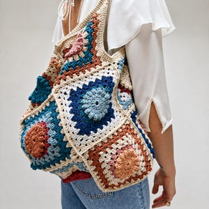 Può includere: Borsa tote all'uncinetto fatta a mano con un design patchwork. La borsa presenta quadrati in vari colori, tra cui blu, beige, arancione e rosa. La borsa è portata a spalla, evidenziando le sue dimensioni e il suo design.