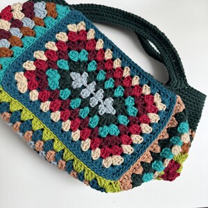 Puede incluir: Un bolso de ganchillo hecho a mano y colorido con un dise&ntilde;o de cuadrados de la abuela. El bolso presenta una variedad de colores, incluyendo turquesa, rojo, verde y beige. La correa es de color verde oscuro. El bolso se utiliza probablemente para llevar objetos.