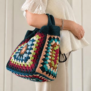 Puede incluir: Un bolso tote de crochet hecho a mano con un dise&ntilde;o de cuadrados de la abuela coloridos. El bolso tiene una base y asas de color verde azulado oscuro, con cuadrados en tonos rojos, azules, verdes y melocot&oacute;n. El bolso es sostenido.