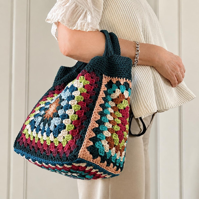 Puede incluir: Un bolso tote de crochet hecho a mano con un dise&ntilde;o de cuadrados de la abuela coloridos. El bolso tiene una base y asas de color verde azulado oscuro, con cuadrados en tonos rojos, azules, verdes y melocot&oacute;n. El bolso es sostenido.