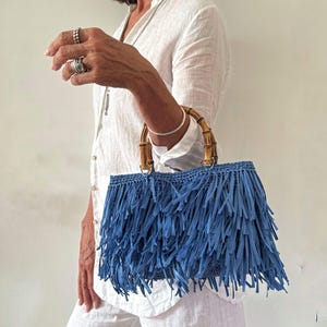 Puede incluir: Bolso azul con flecos y asas de bambú. El bolso es de un tono azul vibrante, con largos detalles de flecos. Las asas son de bambú natural, lo que añade un toque de estilo orgánico. El bolso lo sostiene una persona que lleva una camisa blanca.
