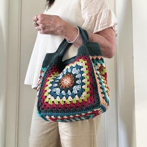 Può includere: Una colorata borsa tote all'uncinetto fatta a mano. La borsa presenta un motivo quadrato con un motivo floreale centrale, utilizzando una varietà di colori tra cui rosso, blu, verde e beige. Ha manici e finiture verde scuro.