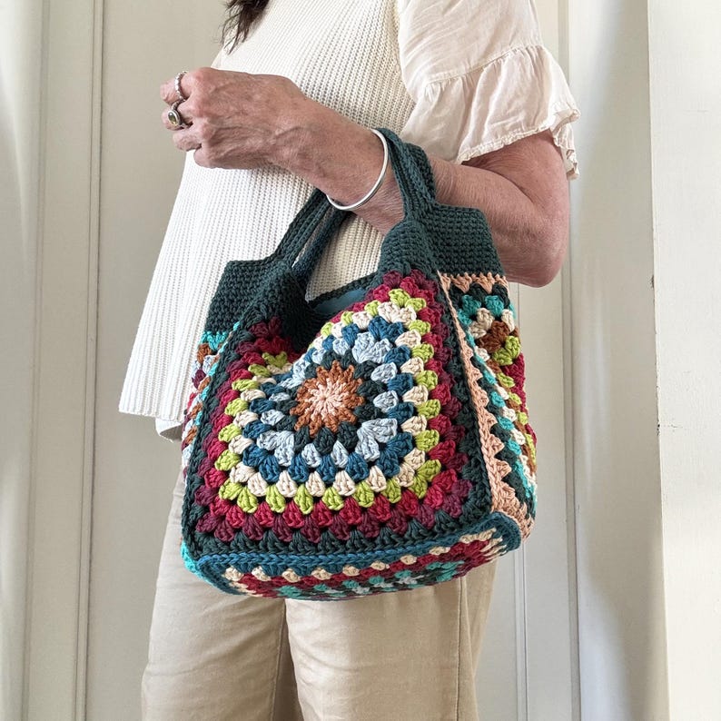 Puede incluir: Un bolso tote de crochet hecho a mano y colorido. El bolso presenta un patr&oacute;n cuadrado con un dise&ntilde;o floral central, utilizando una variedad de colores como rojo, azul, verde y beige. Tiene asas y ribetes de color verde oscuro.
