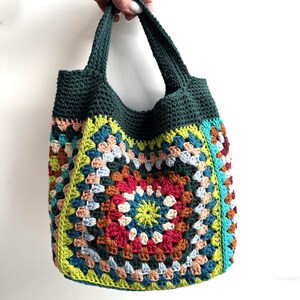 Puede incluir: Bolso tote de crochet hecho a mano con un dise&ntilde;o de cuadrados de la abuela colorido. El bolso presenta una parte superior y asas de color verde oscuro, con un vibrante mosaico de cuadrados rojos, amarillos, azules y marrones. El bolso se sostiene sobre un fondo blanco.