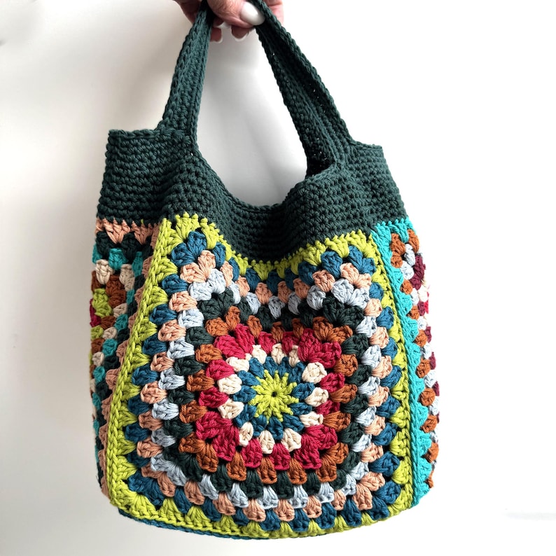 Puede incluir: Bolso tote de crochet hecho a mano con un dise&ntilde;o de cuadrados de la abuela colorido. El bolso presenta una parte superior y asas de color verde oscuro, con un vibrante mosaico de cuadrados rojos, amarillos, azules y marrones. El bolso se sostiene sobre un fondo blanco.