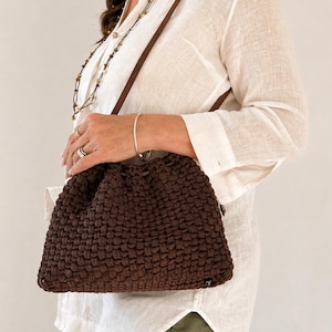Può includere: Una borsa marrone intrecciata con chiusura a coulisse e cinturino in pelle marrone. La borsa è realizzata in un materiale spesso e strutturato e ha una forma rettangolare. La borsa è portata a tracolla.